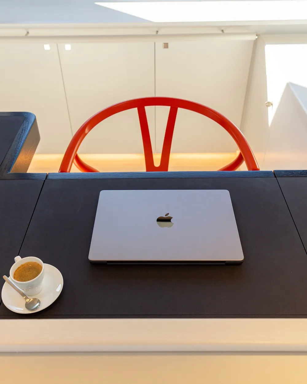 A notebook and an espresso cup on a dark writing desk beside an orange chair