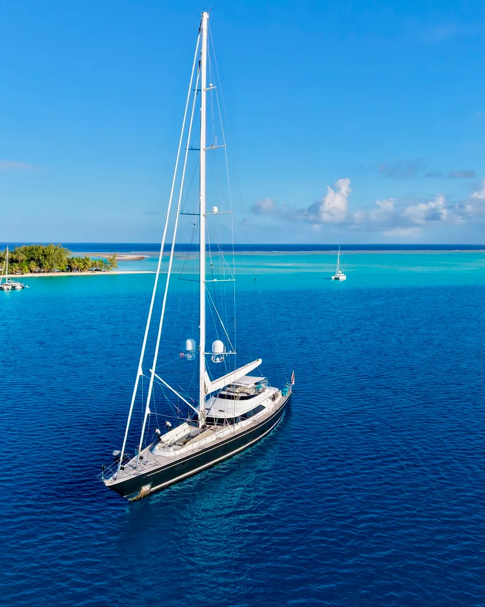 Serendipity I under sail near a Polynesian atoll with a palm-fringed islet and another yacht in the distance