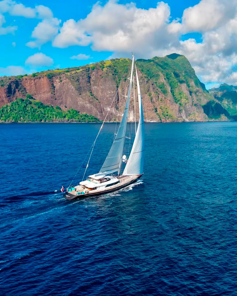 Serendipity I under full sail along the green cliffs of a Polynesian island